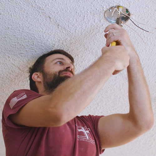 Greysonn Eiland, the owner of Eiland Electric, is shown from the chest up, focused on installing a new light fixture. He is wearing a maroon Eiland Electric t-shirt and is holding a pair of yellow-handled wire strippers, working on the wiring inside a round electrical box mounted on a white, textured ceiling. He is looking up intently at his work, with several wires hanging from the box.