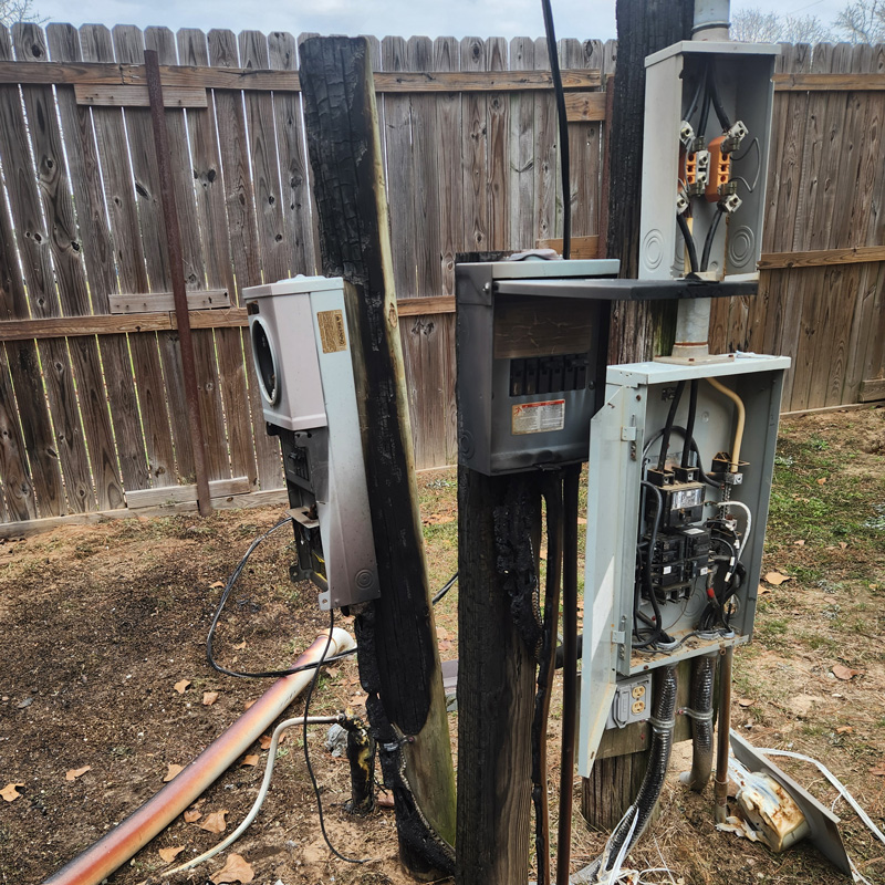 A wooden utility pole, severely charred by fire, stands next to a cluster of damaged electrical boxes. The boxes are open, revealing burnt and exposed wiring and components. The ground around the base of the pole is also scorched, with a broken conduit lying on the dirt. This image represents the type of emergency electrical service calls that Eiland Electric handles, where they are called to repair or replace damaged service equipment after an incident like a fire. A wooden fence is in the background.