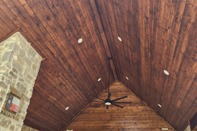 A beautiful outdoor living space features a vaulted, tongue and groove wood ceiling with a dark stain. Recessed lighting is strategically placed throughout the ceiling, illuminating the space. A black ceiling fan with six blades hangs from the center of the peak, providing air circulation. The Eiland Electric logo is visible on a sticker near the fan control box. The walls are constructed of a combination of brown brick and stone, with large windows providing a view of the outdoors.