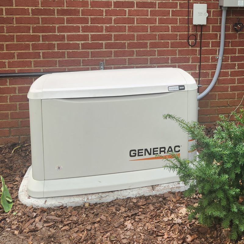 A large, white Generac home backup generator is installed on a concrete pad next to a red brick house. A gray electrical conduit runs down the wall, connecting the generator to the house's electrical system. The surrounding area is covered in mulch and has some green plants. This image illustrates the type of generator installation services offered by Eiland Electric.