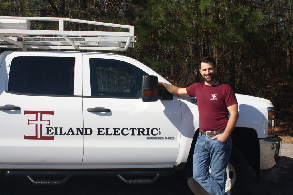 Greysonn Eiland, the owner of Eiland Electric, is standing next to a white company truck. He is smiling at the camera and casually leaning against the front passenger fender with one hand in his pocket. He is wearing a maroon Eiland Electric t-shirt and jeans. The side of the truck clearly displays the Eiland Electric logo and company name. A white ladder rack is visible on the roof. The background is a wooded area.