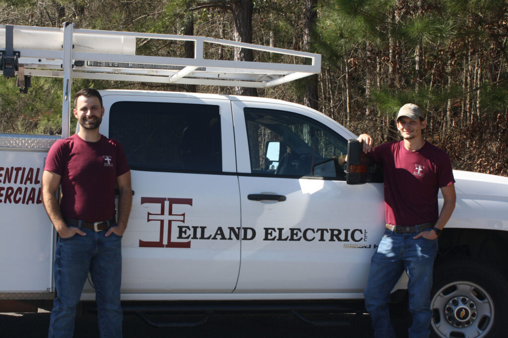 Greysonn Eiland and Bill Ruth are standing next to a white Eiland Electric truck. Both men are wearing matching maroon t-shirts with the Eiland Electric logo. The owner, Greysonn Eiland, is on the left, smiling at the camera with his hands in his pockets. Bill Ruth is on the right, also smiling and leaning on the truck's passenger-side mirror with one hand in his pocket. The truck has the Eiland Electric logo and "RESIDENTIAL COMMERCIAL" text on the door, and a white ladder rack is mounted on top. The background consists of a wooded area with trees and bushes.