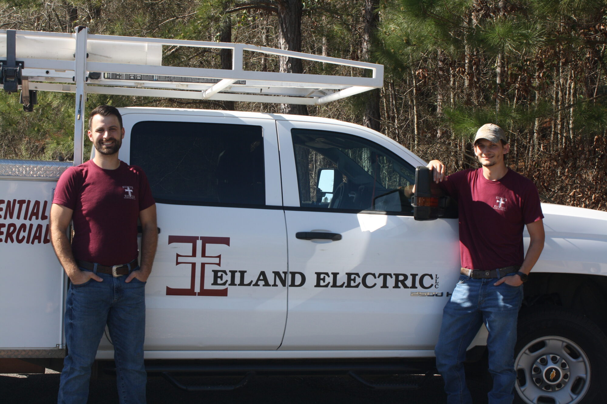 Greysonn Eiland and Bill Ruth are standing next to a white Eiland Electric truck. Both men are wearing matching maroon t-shirts with the Eiland Electric logo. The owner, Greysonn Eiland, is on the left, smiling at the camera with his hands in his pockets. Bill Ruth is on the right, also smiling and leaning on the truck's passenger-side mirror with one hand in his pocket. The truck has the Eiland Electric logo and "RESIDENTIAL COMMERCIAL" text on the door, and a white ladder rack is mounted on top. The background consists of a wooded area with trees and bushes.